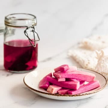 A plate of bright pink Middle Eastern Pickled Turnips sits next to a jar of vibrant pink pickling liquid on a white surface.