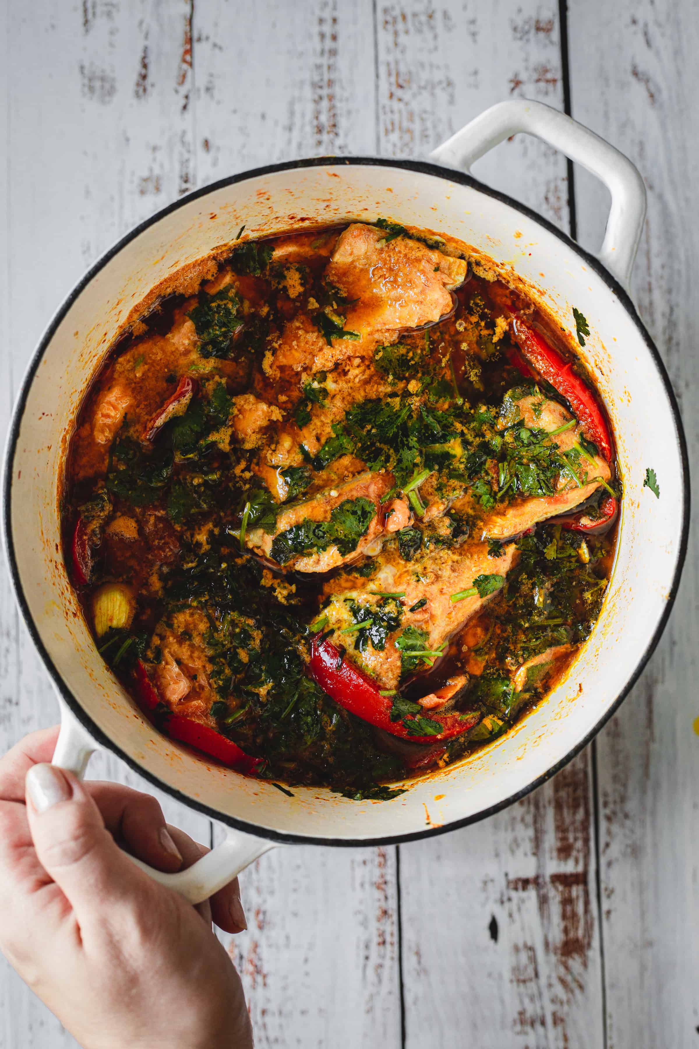 A hand holds a white pot filled with Moroccan salmon stew, herbs, and red peppers on a rustic wooden table.