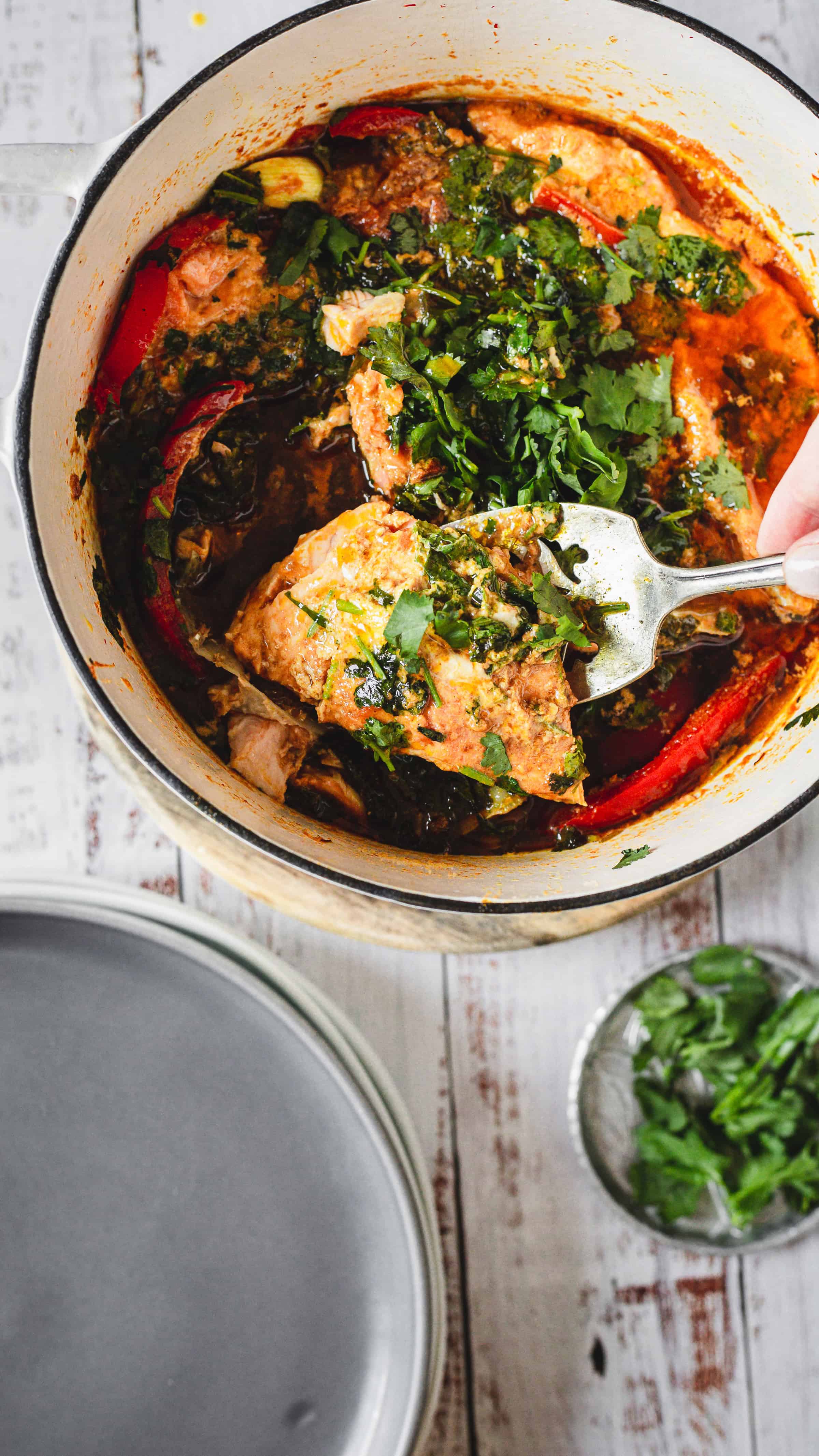 A fork lifts a piece of Moroccan salmon in a red, herb-filled stew from a white pot on a rustic table.