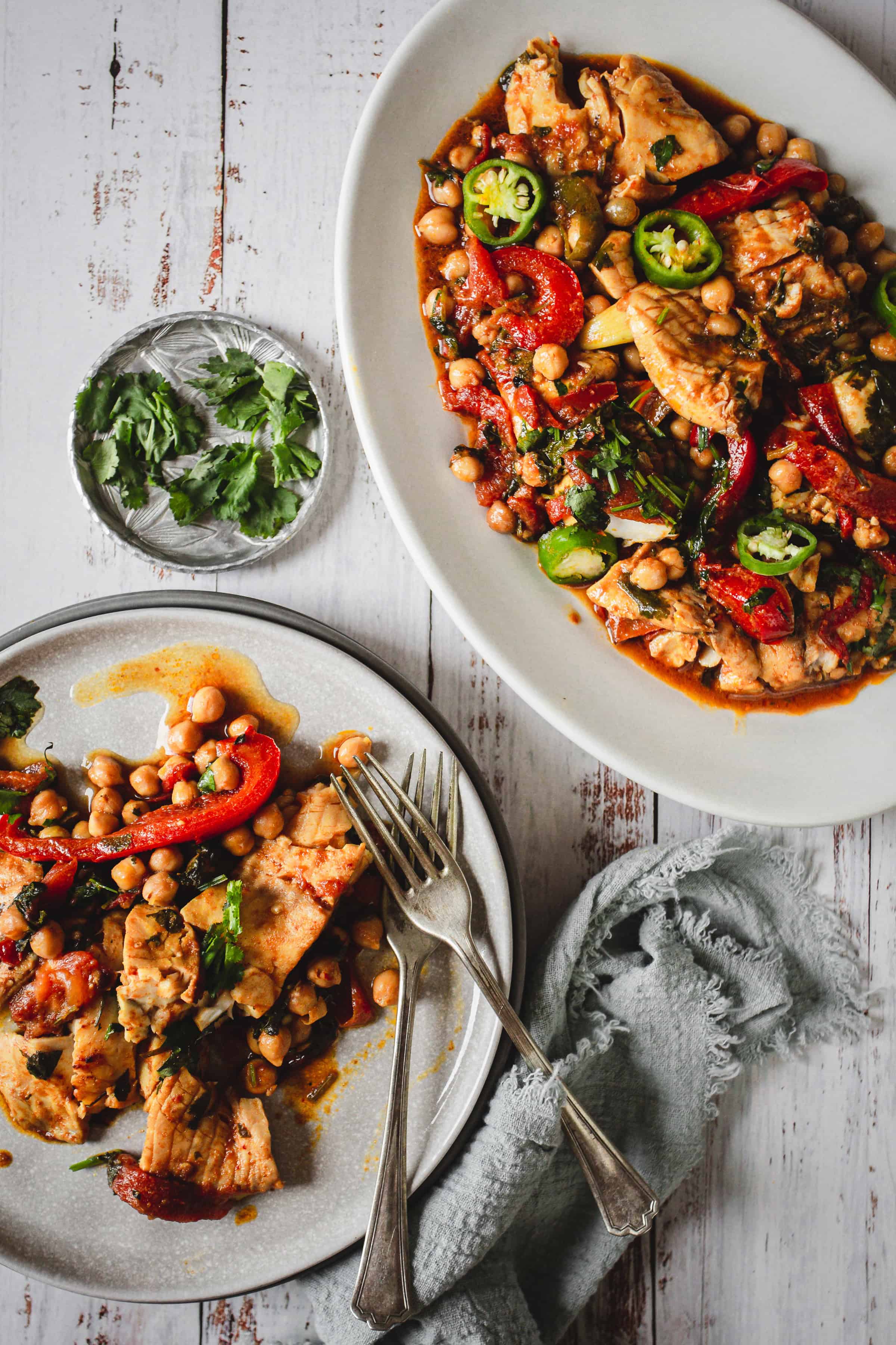 A plate and platter of Moroccan chicken stew with chickpeas, peppers, and herbs sits alongside savory Moroccan salmon, served with forks and a napkin.