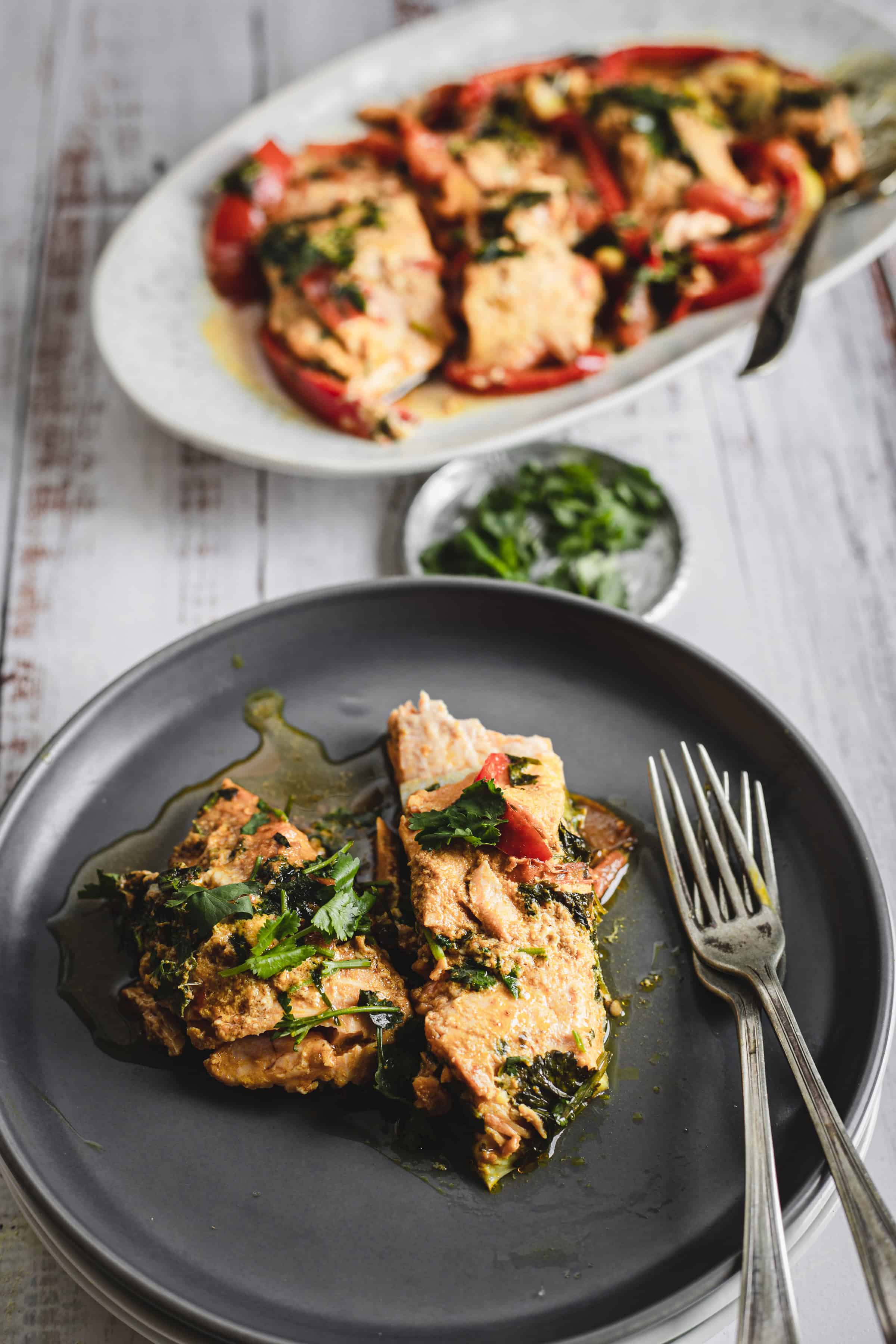 Two servings of Moroccan salmon with herbs and sauce on a black plate, with more on a platter in the background.