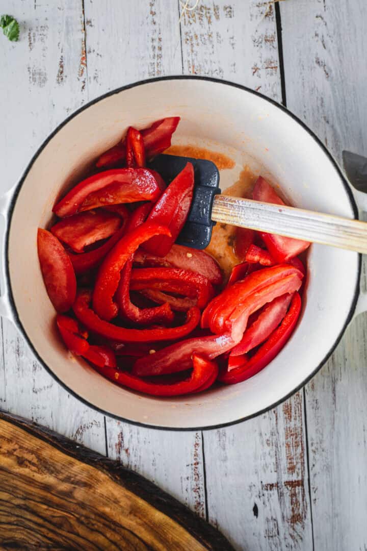 Red bell pepper slices being stirred in a white pot with a black spatula on a rustic white wooden surface, perfect for adding to Moroccan salmon.