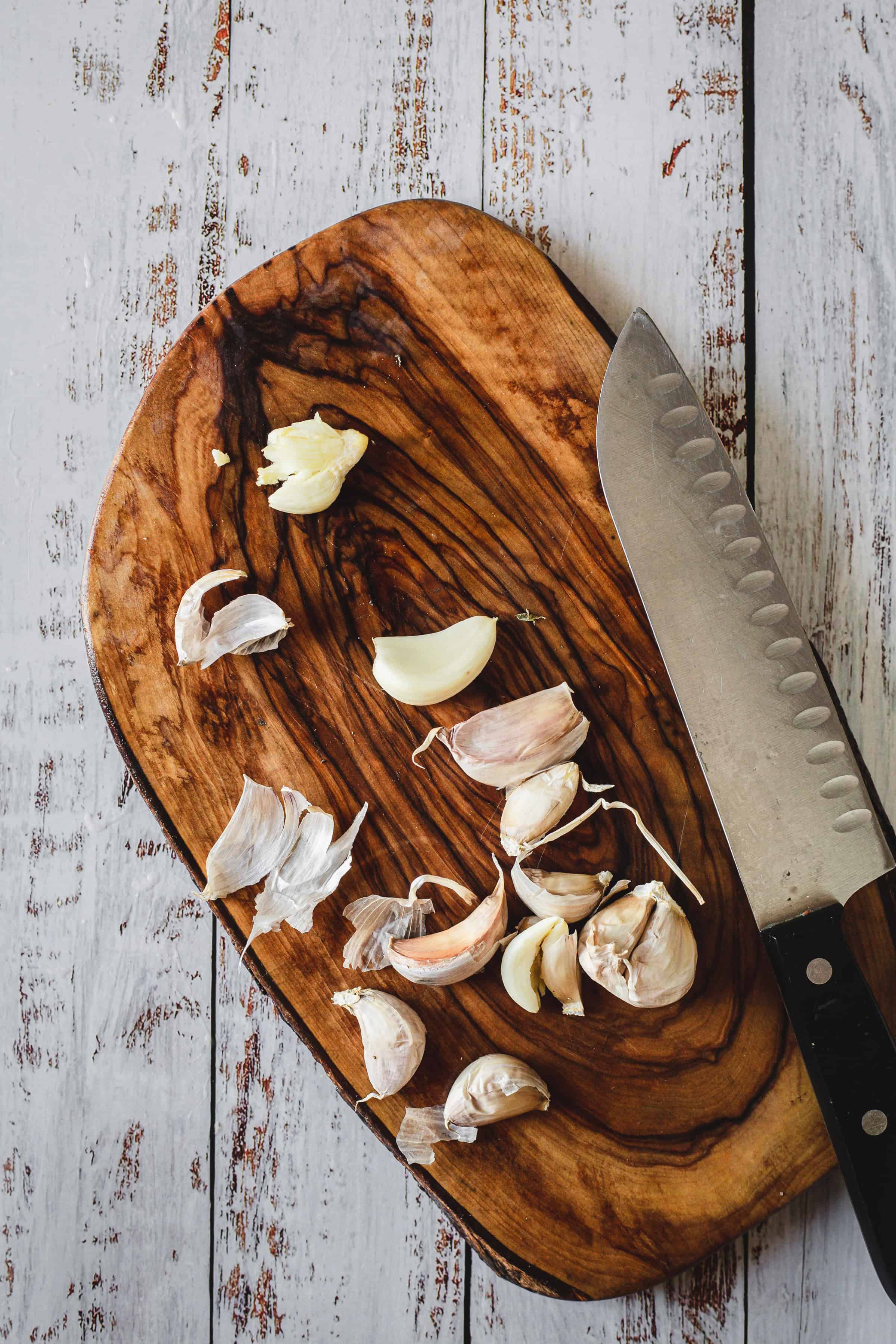 Garlic cloves and a kitchen knife on a wooden cutting board, set on a rustic white table&mdash;perfect prep for Moroccan salmon.