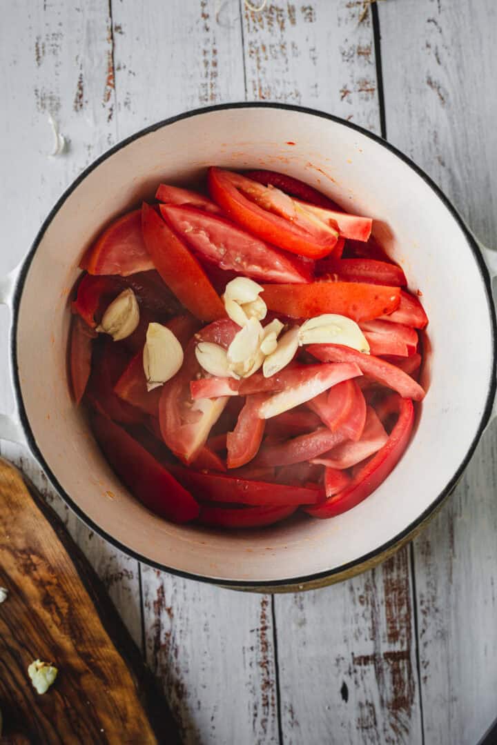 Sliced tomatoes and garlic cloves in a white pot on a rustic white wooden table, perfect for preparing Moroccan salmon.