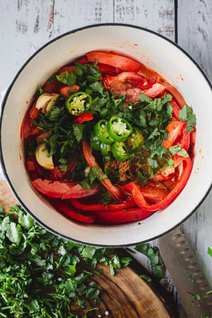 Chopped peppers, tomatoes, herbs, and spices simmer in a pot for Moroccan salmon, with fresh cilantro and a knife on a cutting board nearby.
