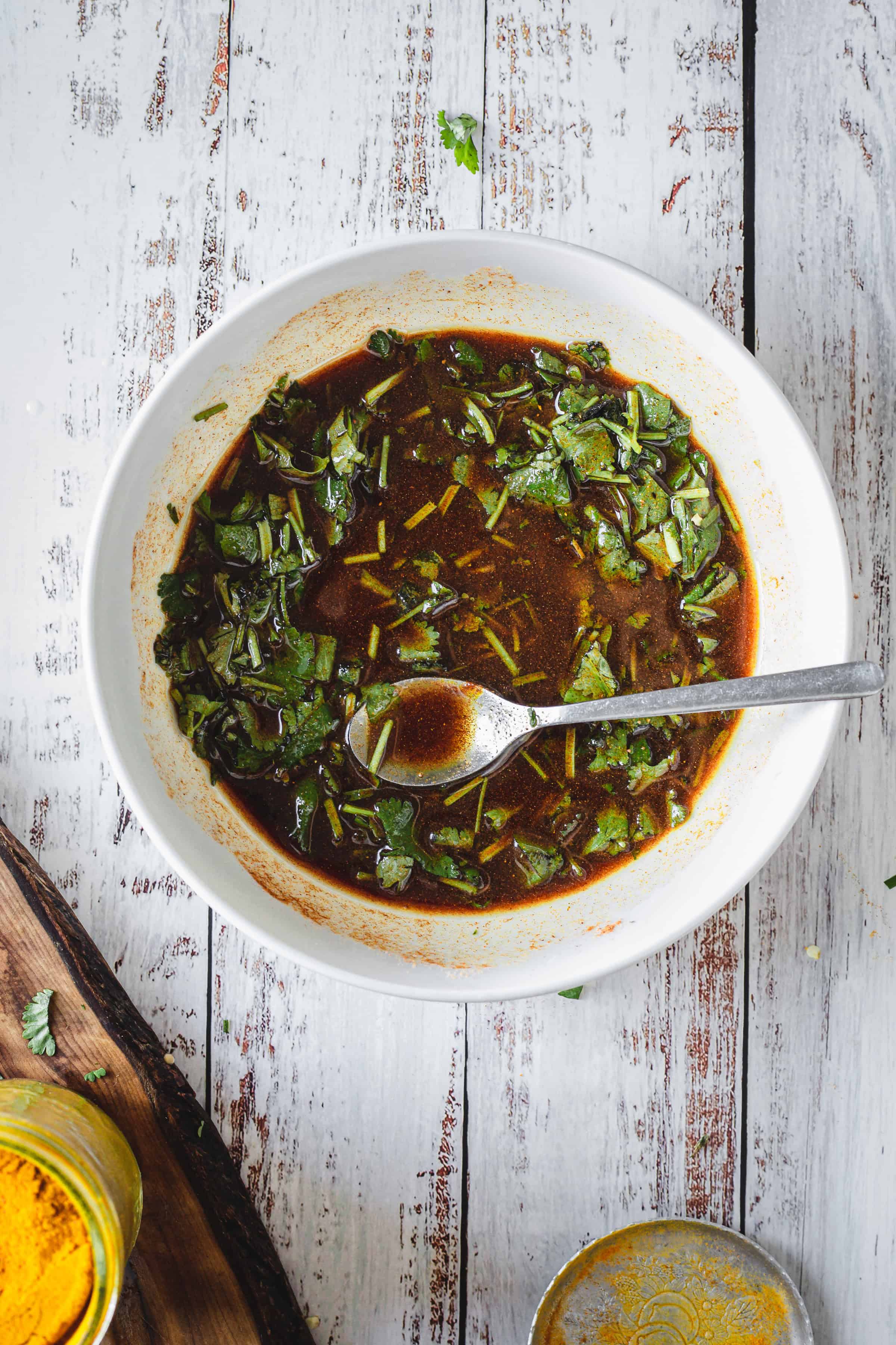 A bowl of dark dressing with chopped herbs&mdash;perfect for Moroccan salmon&mdash;sits with a spoon on a rustic white wooden surface.