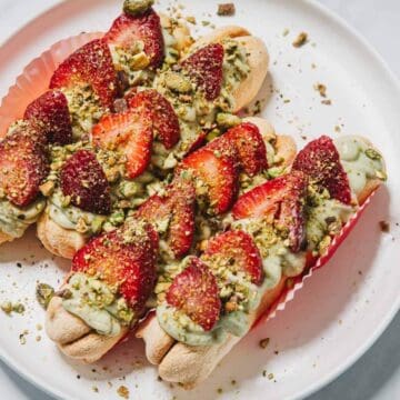 Three ladyfinger desserts topped with green cream, sliced strawberries, and chopped pistachios on a white plate.