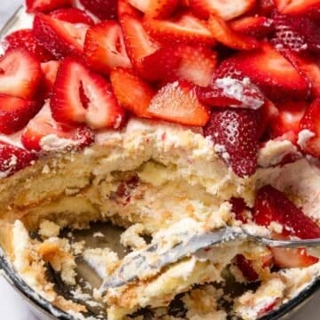 A dish of strawberry tiramisu with a spoon and a portion already served, topped with fresh sliced strawberries.