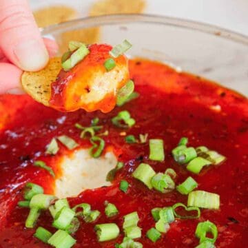 A hand dips a round chip into a cheesy dip topped with red sauce and chopped green onions.
