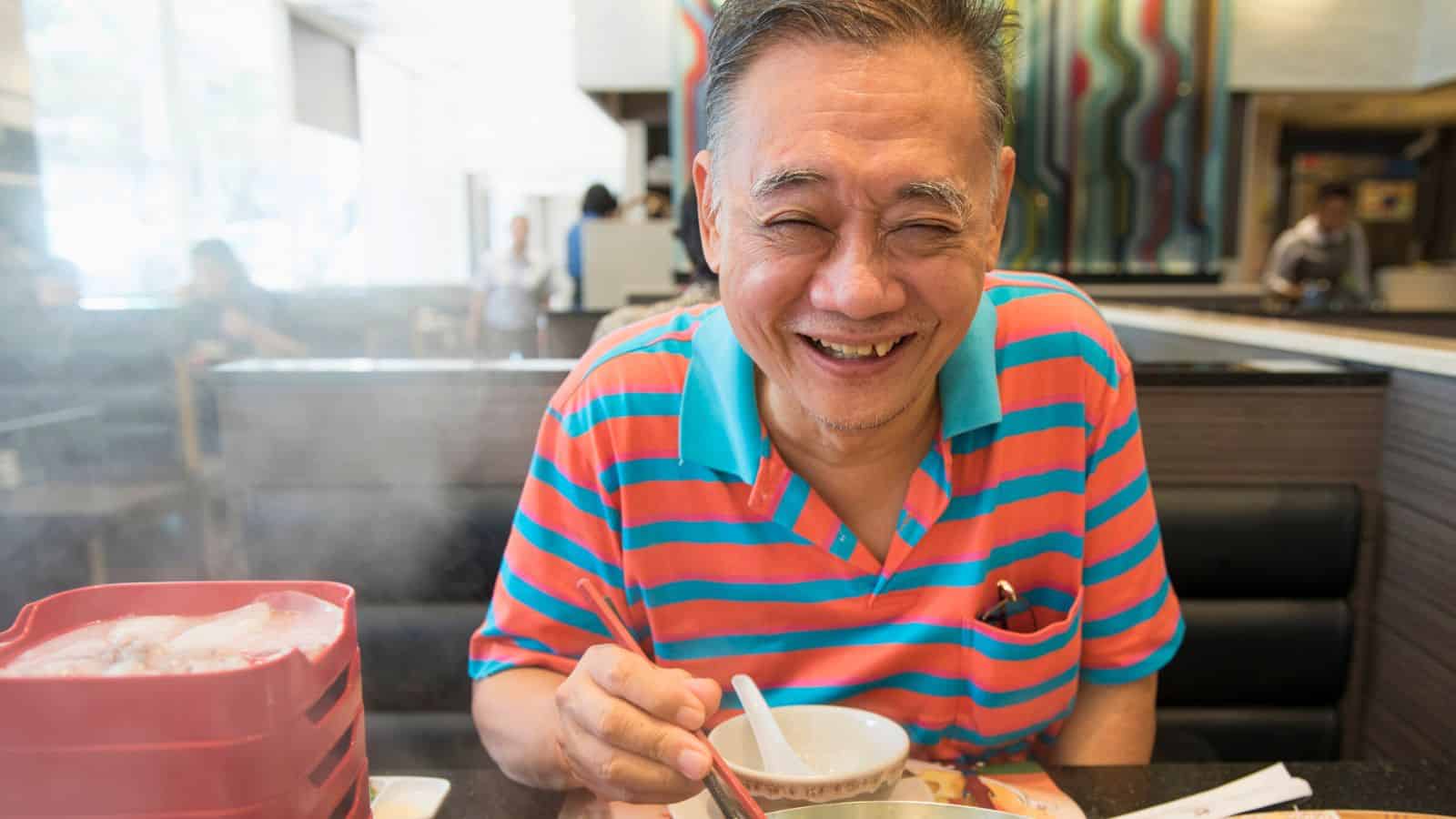 Smiling man in a striped shirt enjoying a meal at a restaurant with a bowl and chopsticks.