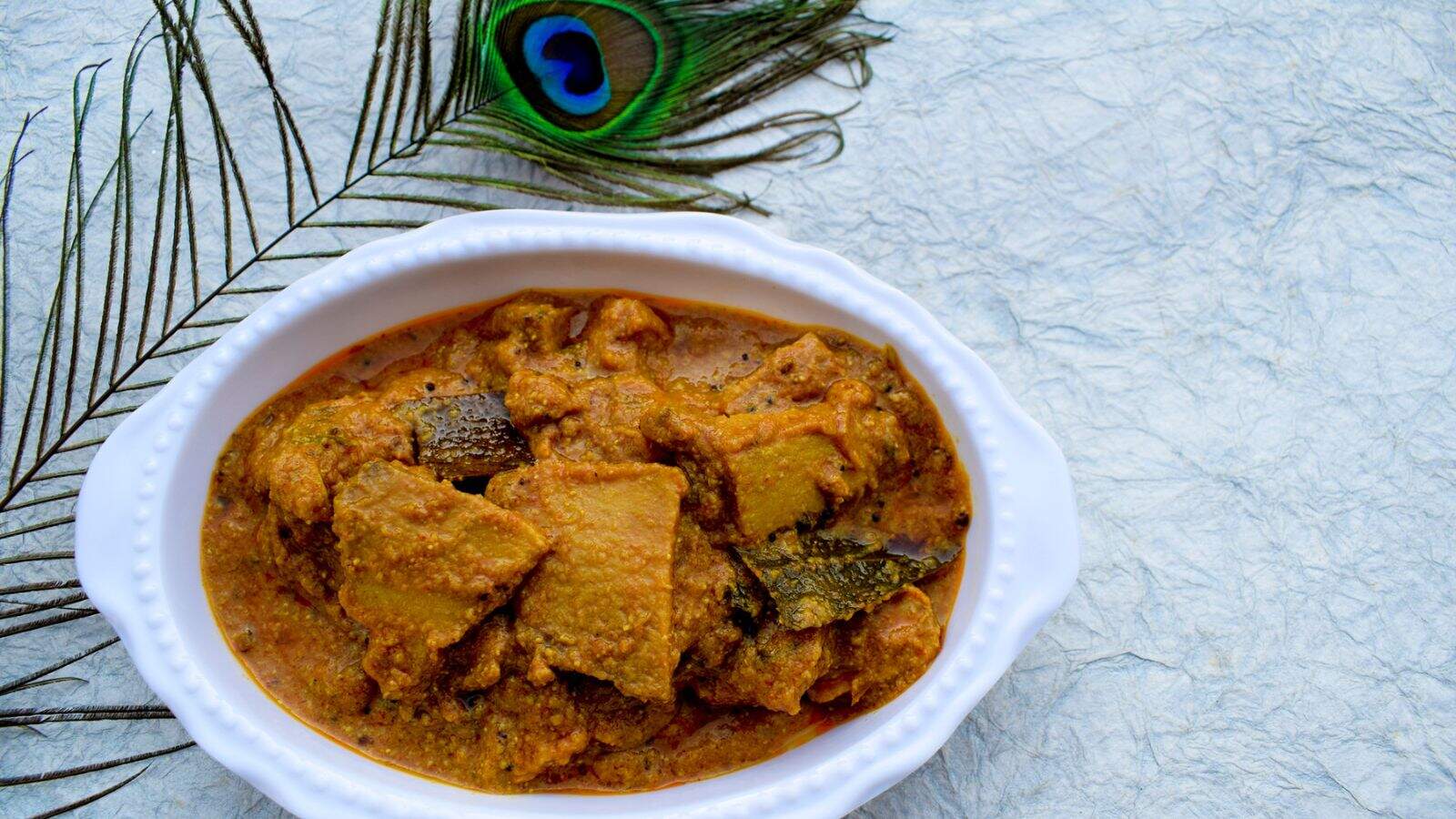 A white oval dish filled with a thick, orange-brown curry containing pieces of vegetables or meat. A peacock feather is placed beside the dish on a light textured surface.