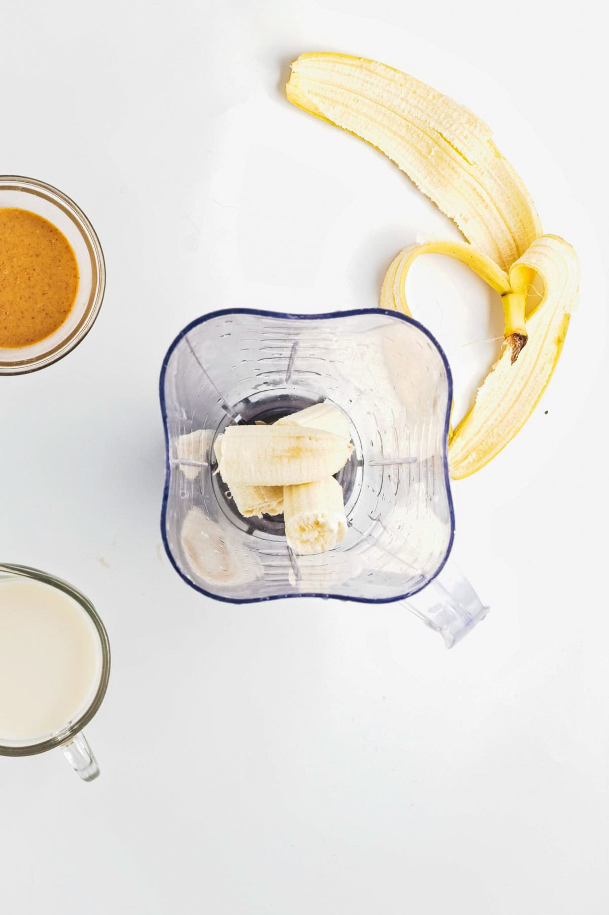 A blender with banana pieces, a peeled banana, a glass of milk, and a cup of nut butter on a white surface-perfect ingredients for Peanut Butter Banana Overnight Chia Oats.