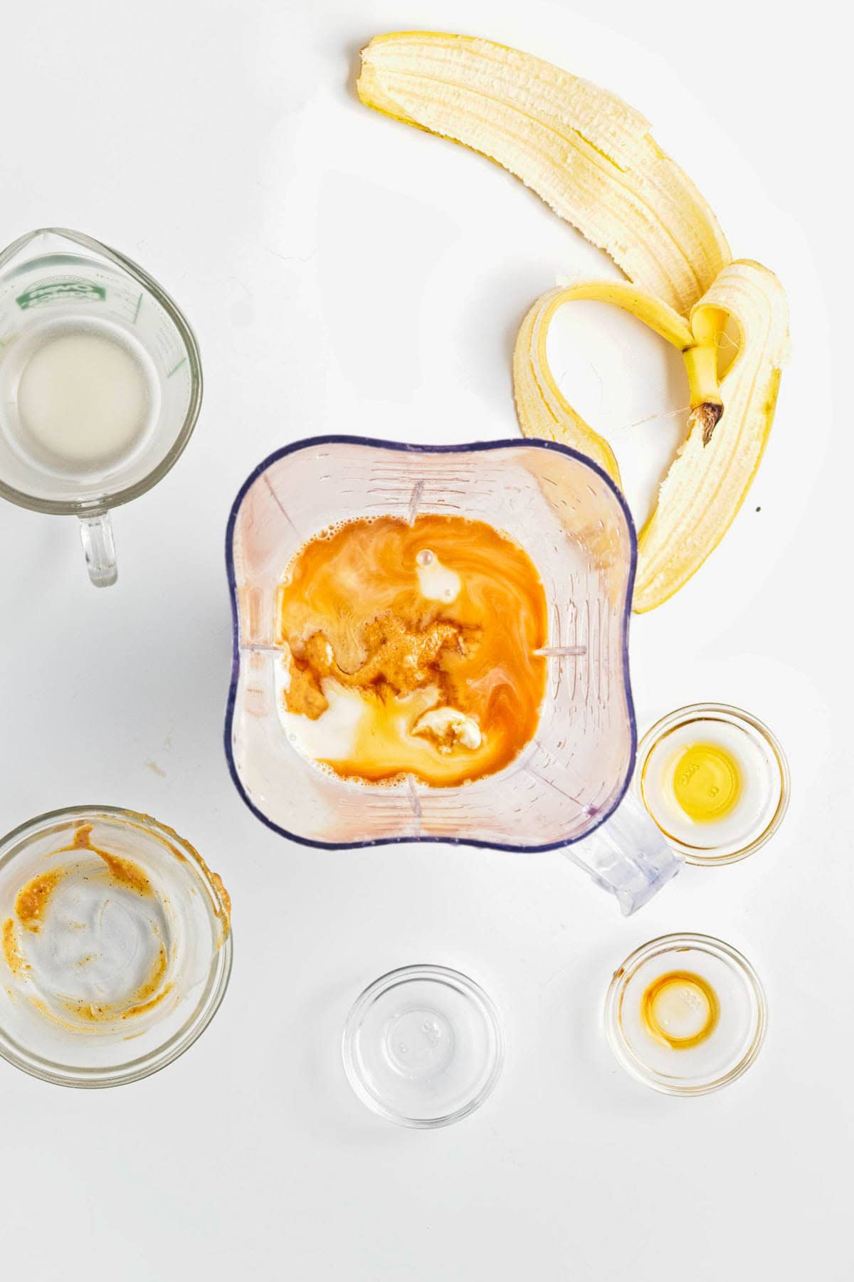 Ingredients for a smoothie in and around a blender: banana, nut butter, milk, and flavorings on a white surface-perfect for whipping up Peanut Butter Banana Overnight Chia Oats.