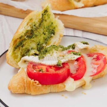 A Pesto Tomato Brie Croissant Sandwich with melted cheese, fresh tomato slices, and green pesto served on a plate.