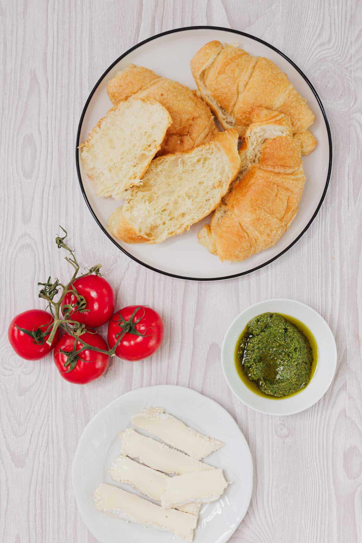 Sliced croissants, vine tomatoes, a bowl of pesto, and cheese slices on a light wooden table—perfect ingredients for a delicious Pesto Tomato Brie Croissant Sandwich.