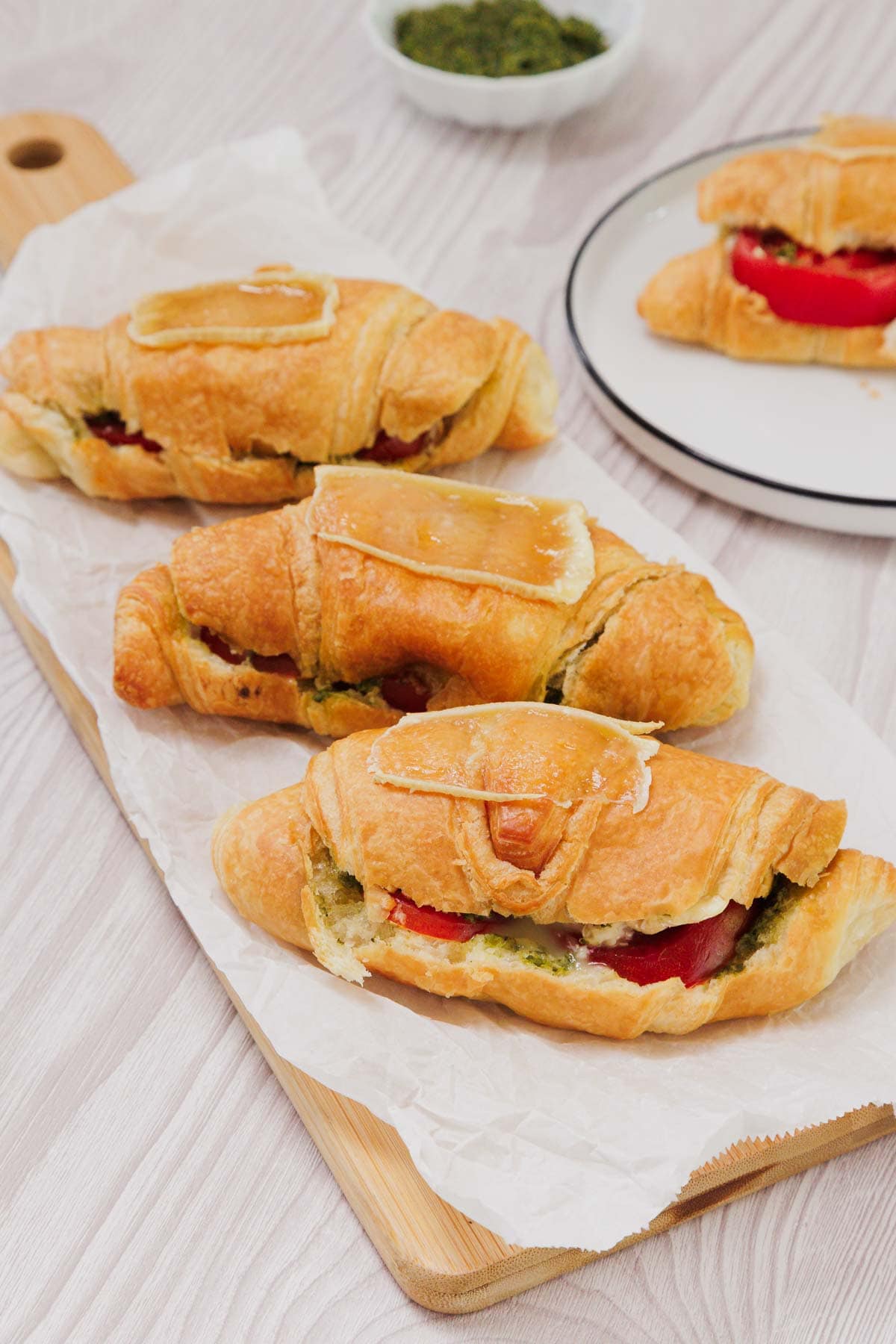 Three Pesto Tomato Brie Croissant Sandwiches with cheese and tomato sit on parchment paper atop a rustic wooden board.