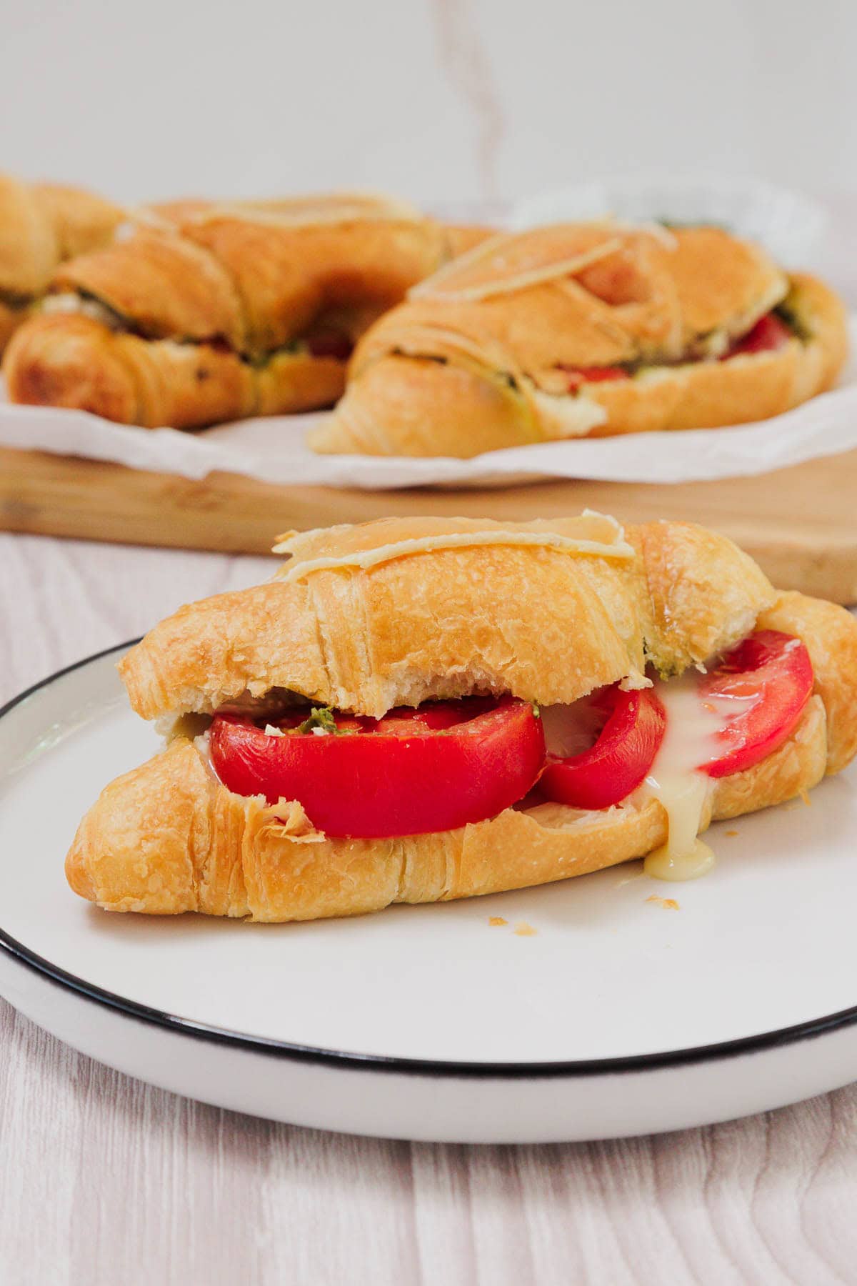 A Pesto Tomato Brie Croissant Sandwich with tomato and melted cheese sits on a plate, with more sandwiches in the background.