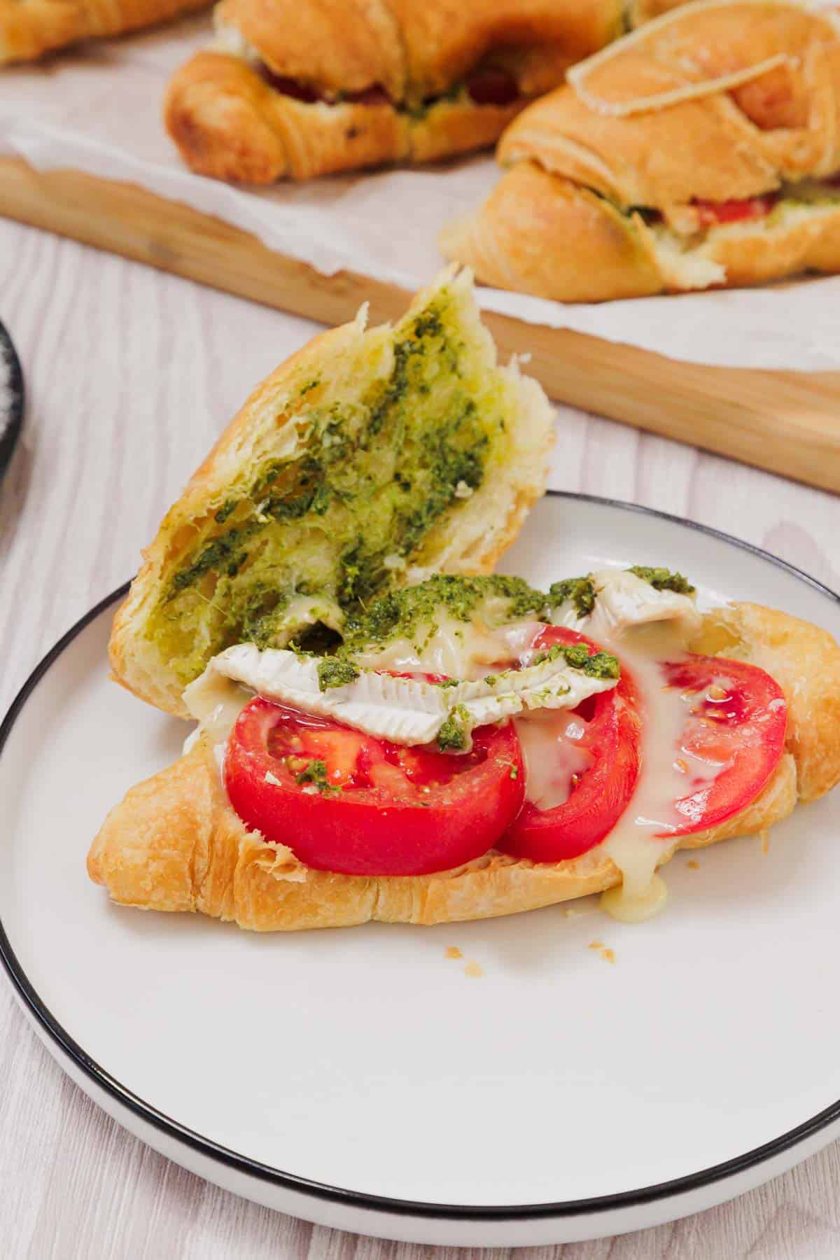 Enjoy a Pesto Tomato Brie Croissant Sandwich, featuring sliced tomato, creamy brie, and green pesto melted inside a flaky croissant on a white plate.