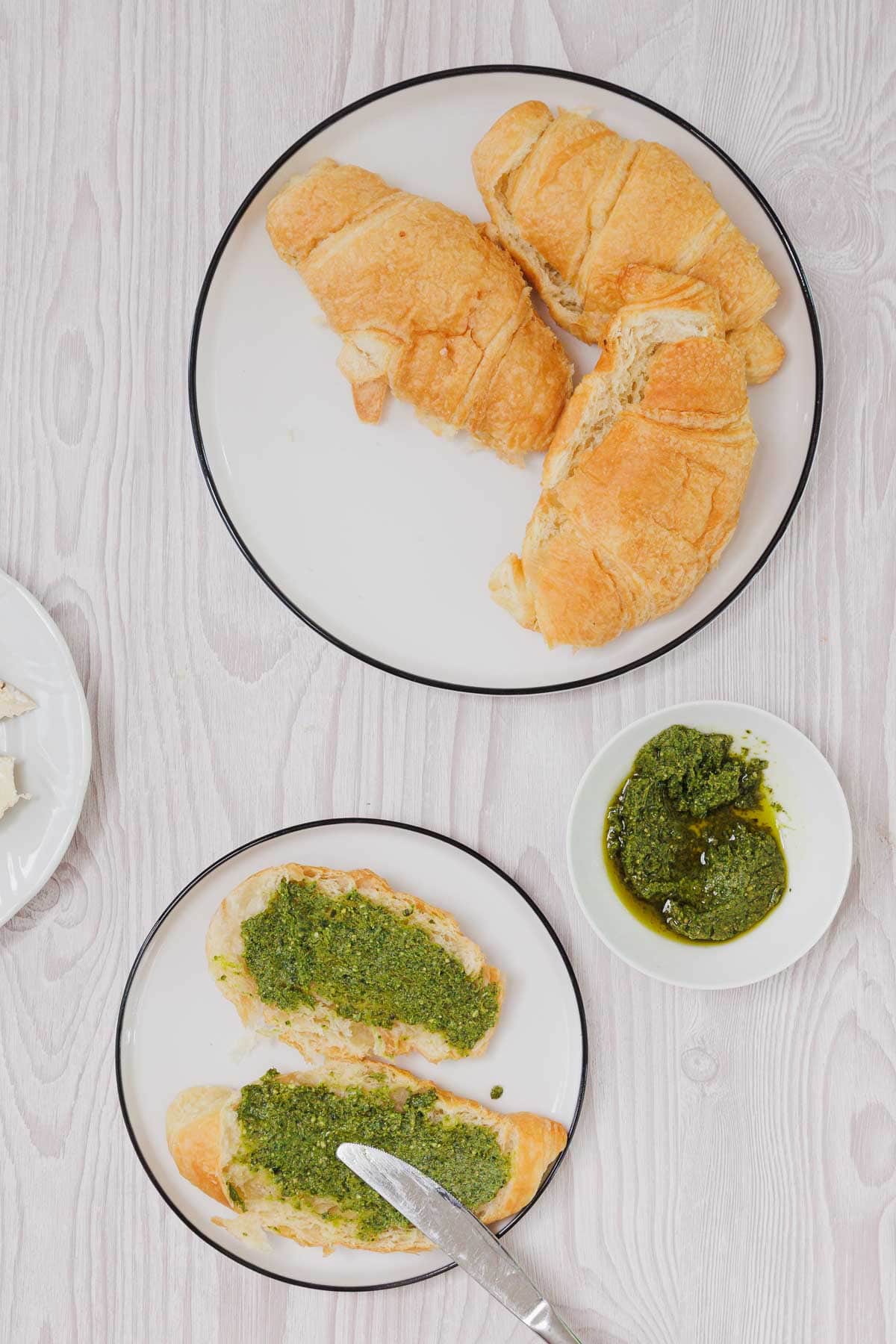 Two croissants on one plate, and two croissant halves with green pesto on another plate suggest the makings of a delicious Pesto Tomato Brie Croissant Sandwich, served alongside a bowl of pesto for extra flavor.