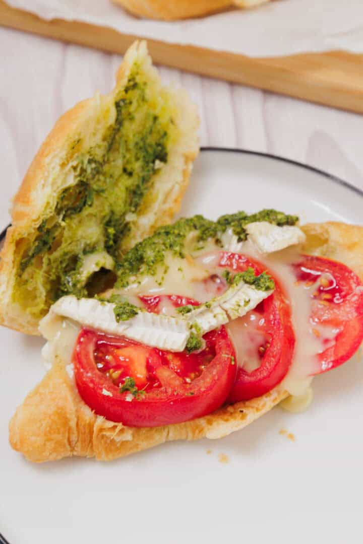 Enjoy a Pesto Tomato Brie Croissant Sandwich, featuring tomato slices, creamy brie cheese, and vibrant green pesto on a flaky croissant, served on a white plate.