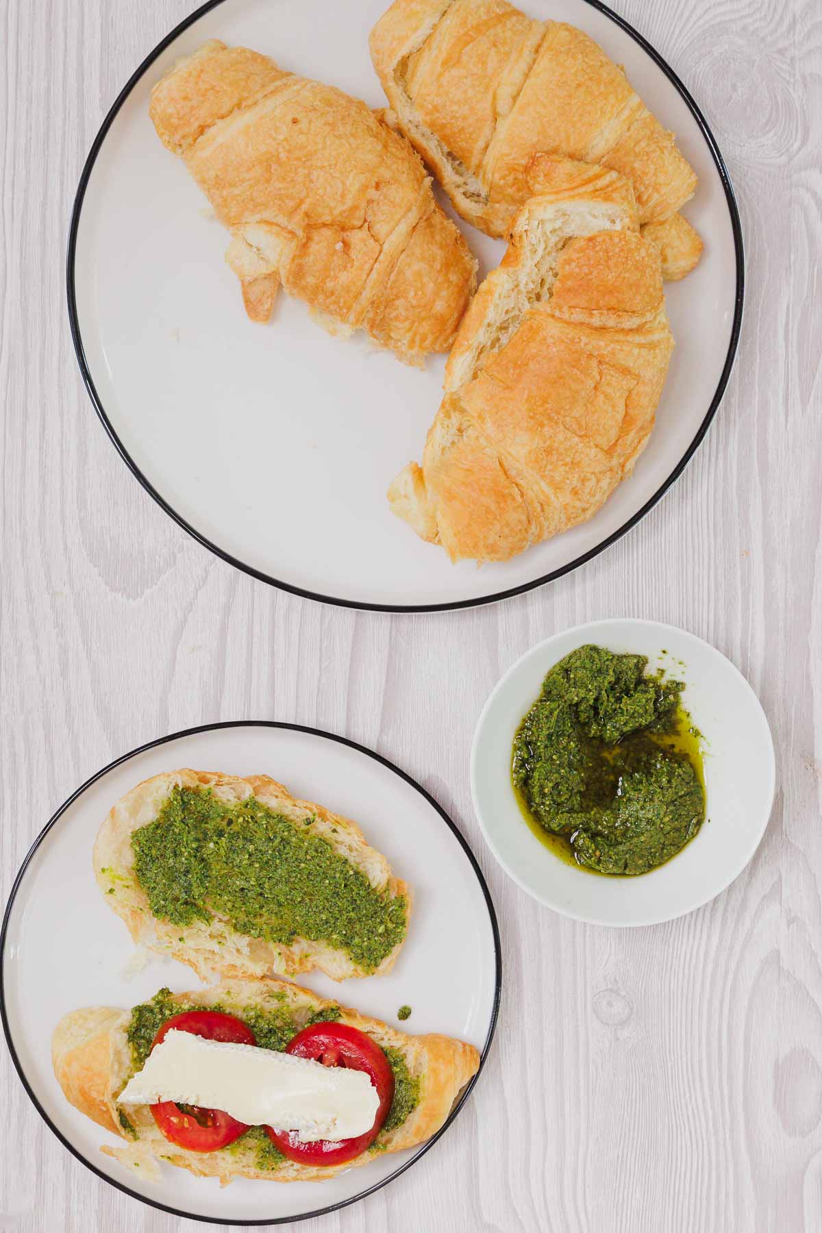Two croissants on one plate, and an open-faced Pesto Tomato Brie Croissant Sandwich with melted cheese on another plate.