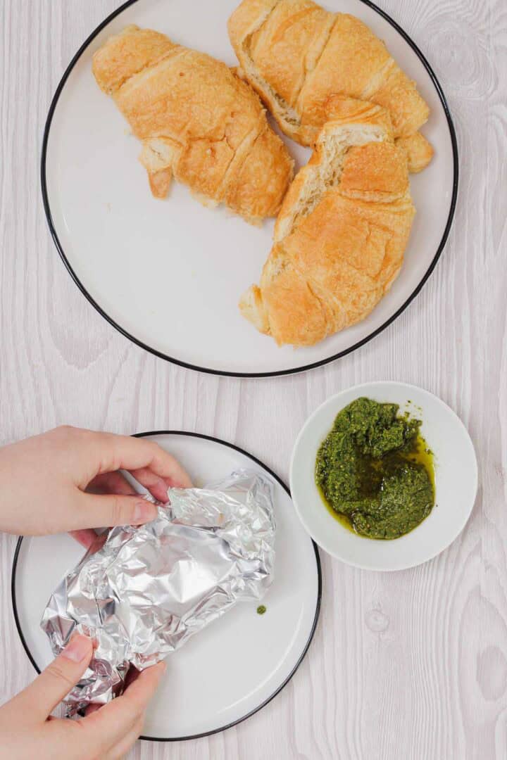 Two Pesto Tomato Brie Croissant Sandwiches on a plate, pesto in a bowl, and hands unwrapping foil on a separate plate.