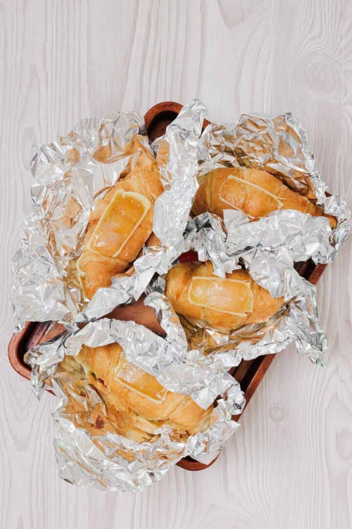 Four baked bread rolls wrapped in crinkled aluminum foil on a wooden tray, resembling a rustic pesto tomato brie croissant sandwich spread, set on a light wooden surface.