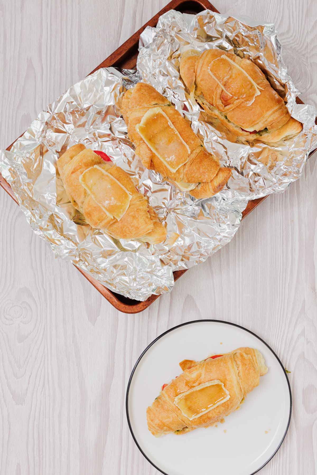 Three Pesto Tomato Brie Croissant Sandwiches with cheese rest on a foil-lined tray, with one on a plate, all set atop a light wooden surface.