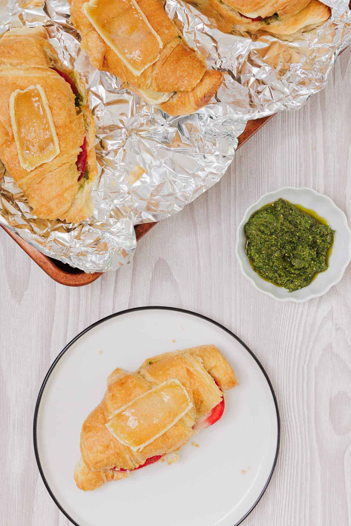 A Pesto Tomato Brie Croissant Sandwich sits on a plate, its flaky layers filled with vibrant pesto, tomato, and brie, while more golden croissants rest on a foil-lined tray nearby.