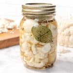 A glass jar of pickled cauliflower with a bay leaf and spices, on a marble counter near a cutting board.