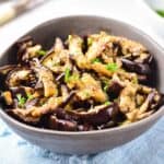 A bowl of pickled eggplant slices garnished with chopped parsley sits on a light blue cloth.