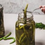 A hand lifts quick pickled green beans from a glass jar with a fork on a light surface.
