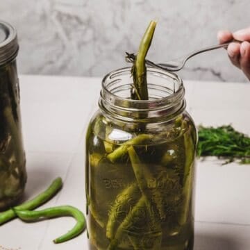 A hand lifts quick pickled green beans from a glass jar with a fork on a light surface.