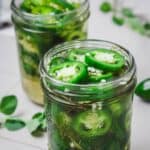 Two glass jars filled with sliced jalapeños in brine, surrounded by green herbs on a light surface.