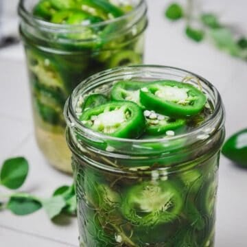Two glass jars filled with sliced jalapeños in brine, surrounded by green herbs on a light surface.