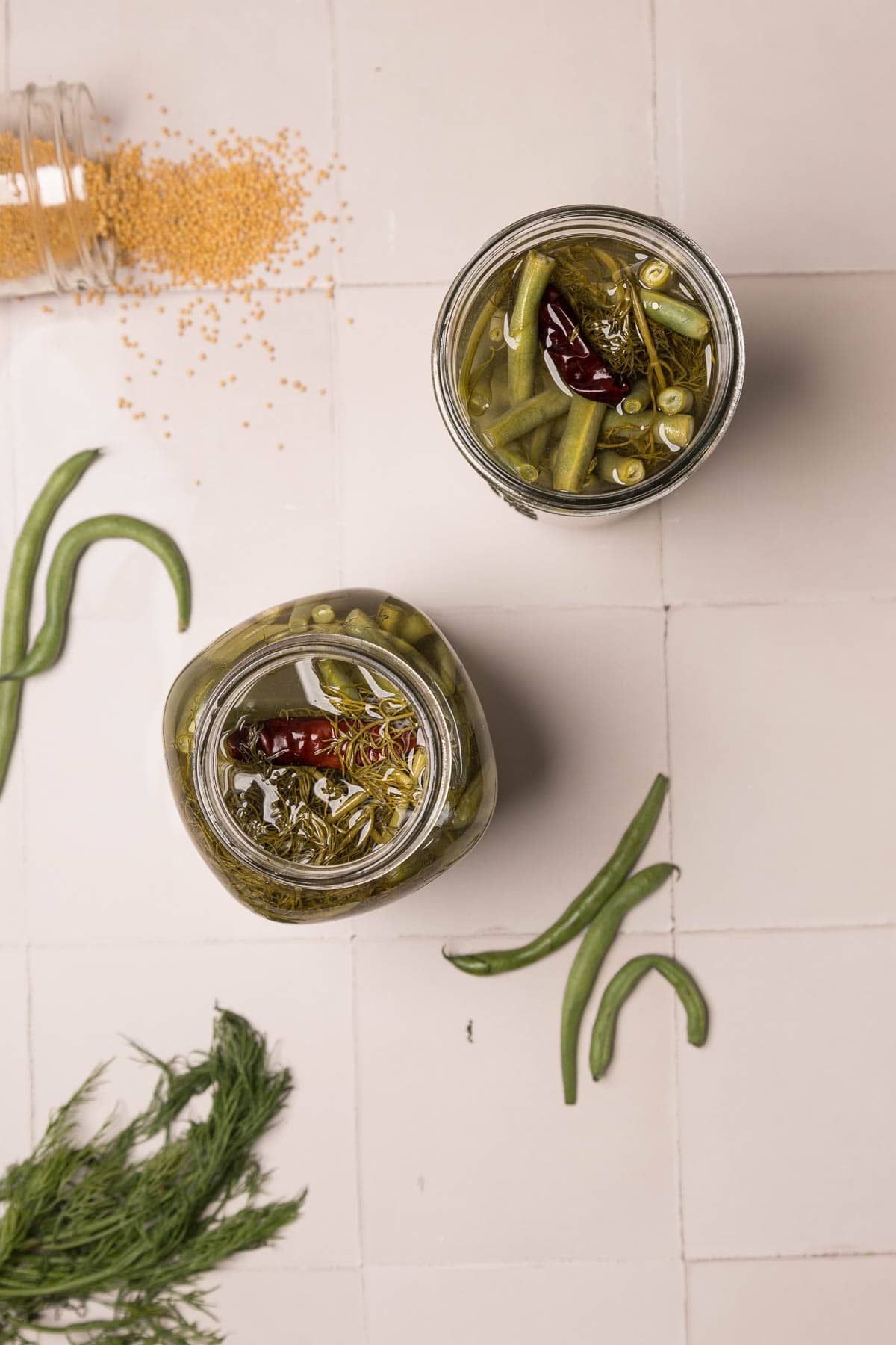 Two jars of pickled green beans with spices on a white tiled surface, with scattered beans, herbs, and mustard seeds.