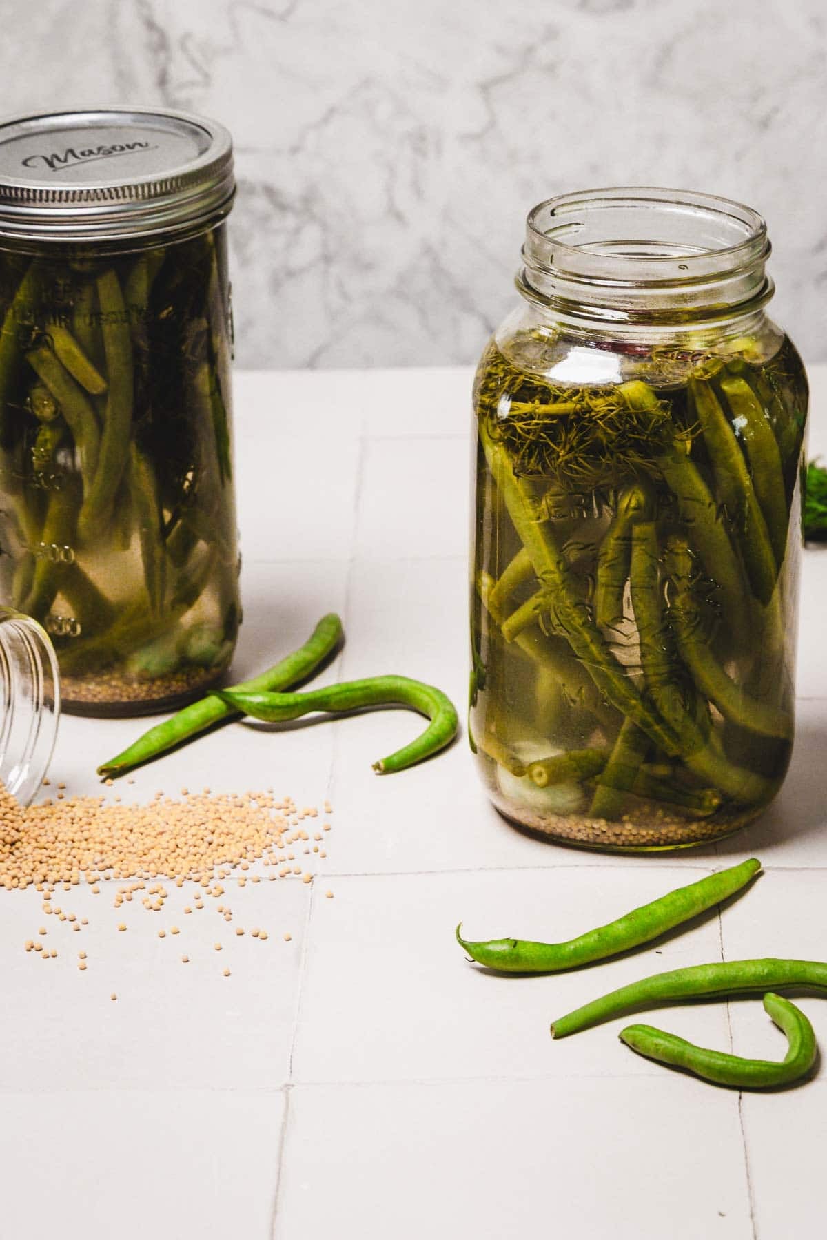 Two jars of pickled green beans with loose green beans and mustard seeds on a white tiled surface.
