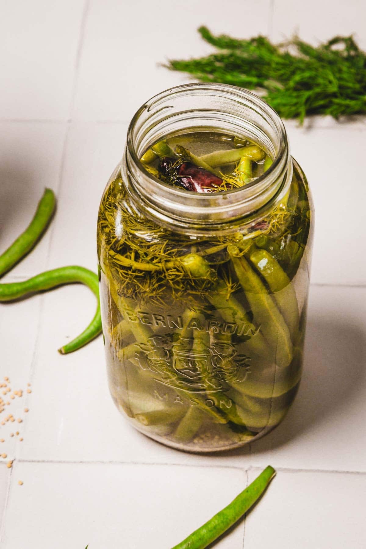 A mason jar filled with pickled green beans, dill, and spices on a white tiled surface.