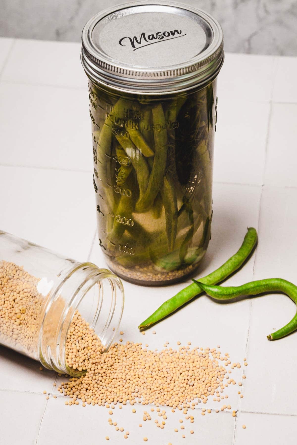 A mason jar of pickled green beans beside spilled mustard seeds and fresh green beans on a white surface.