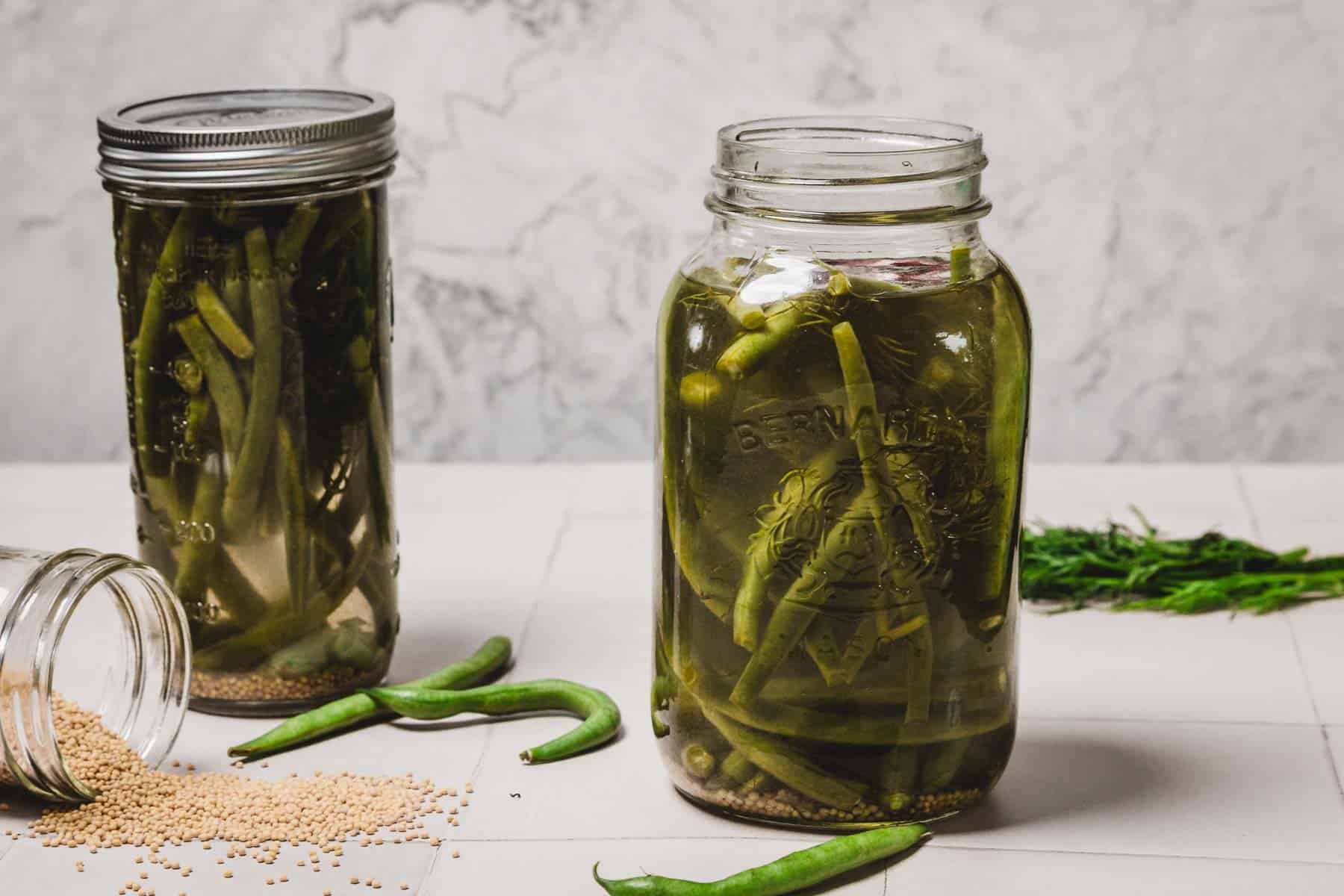 Two jars of pickled green beans, with loose beans, mustard seeds, and dill on a white tile surface.