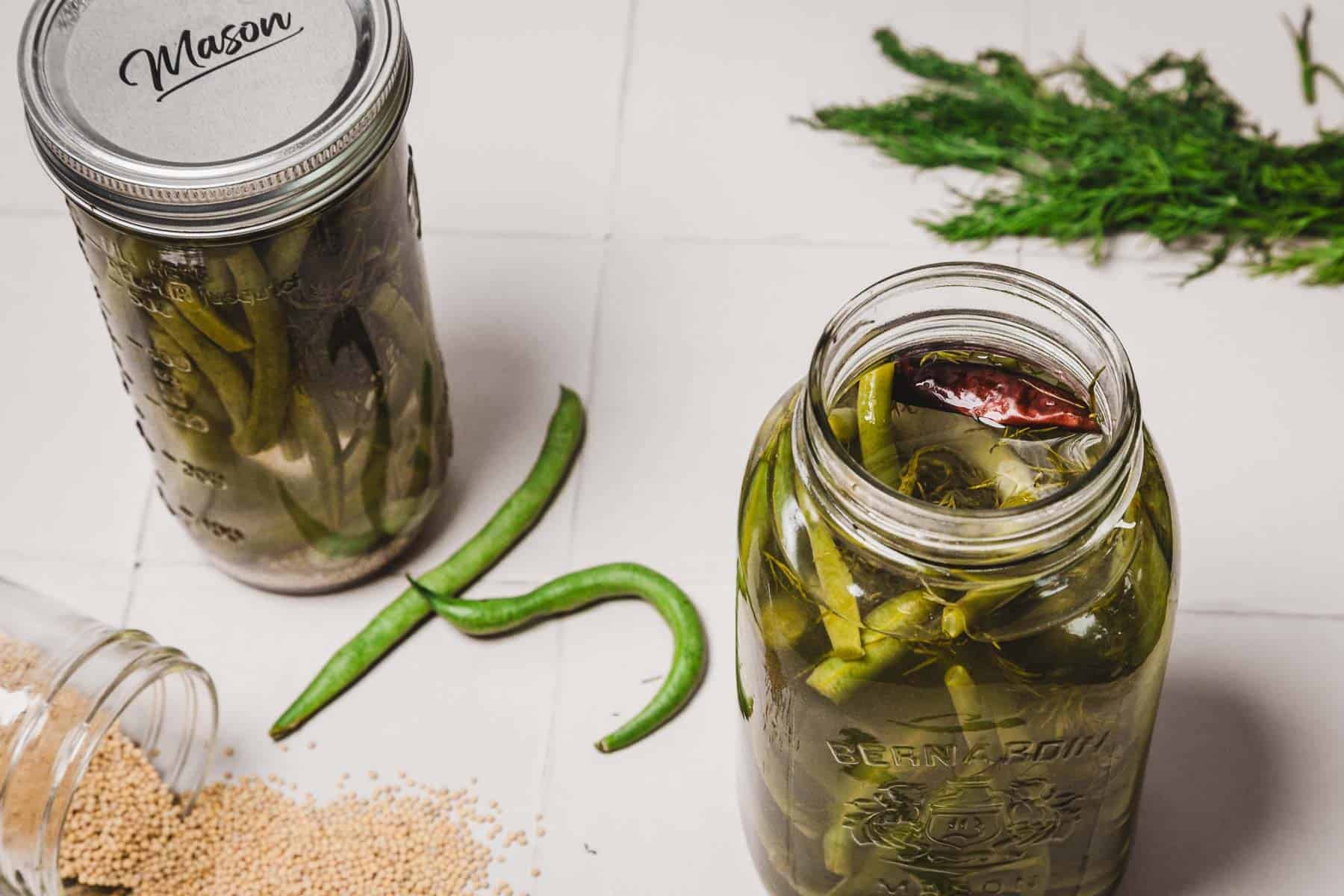 Two jars of pickled green beans, fresh dill, spices, and green beans on a white tiled surface.