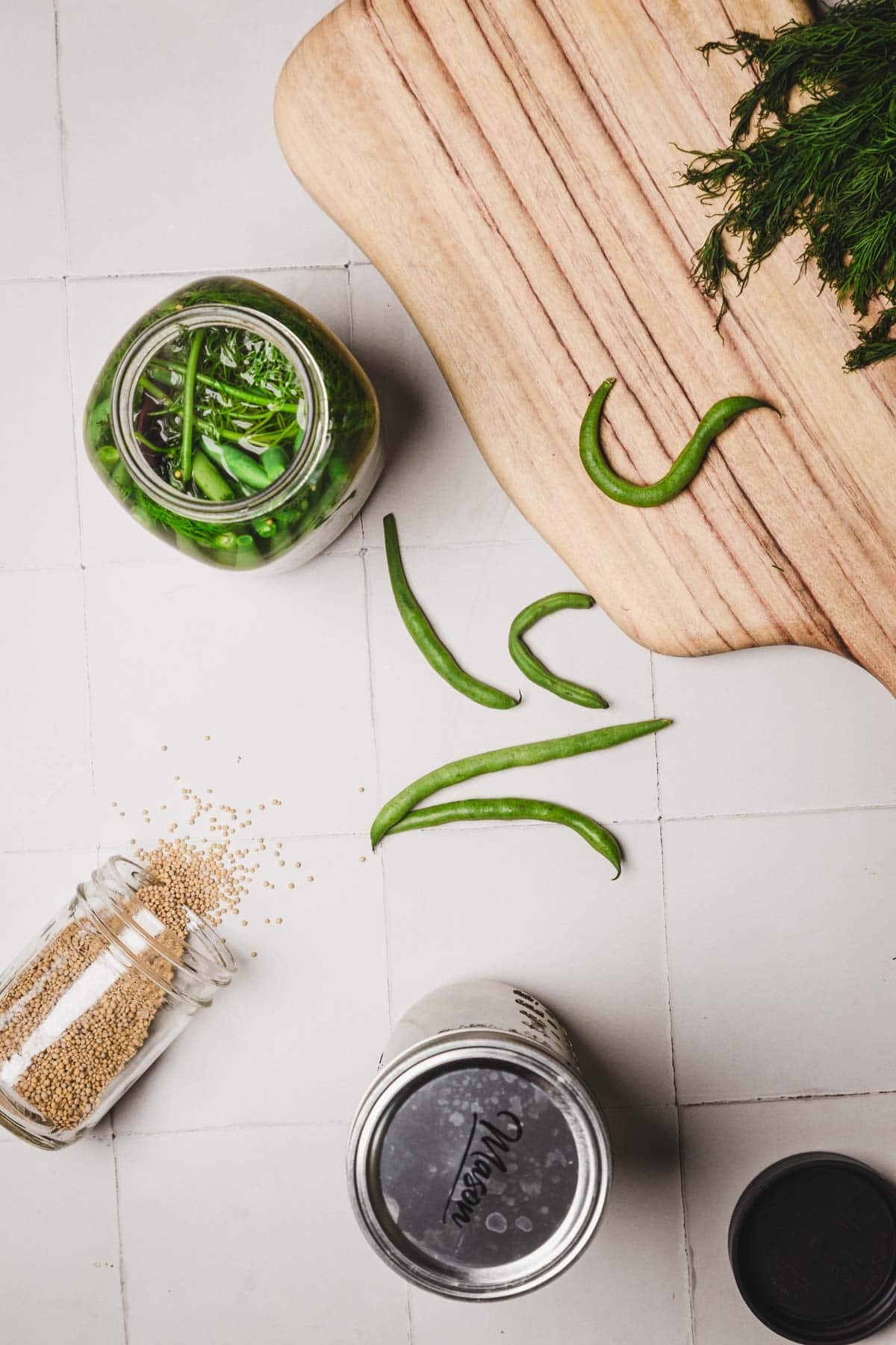 Green beans, fresh dill, and mustard seeds on a tiled surface with a cutting board and a jar of pickling vegetables.