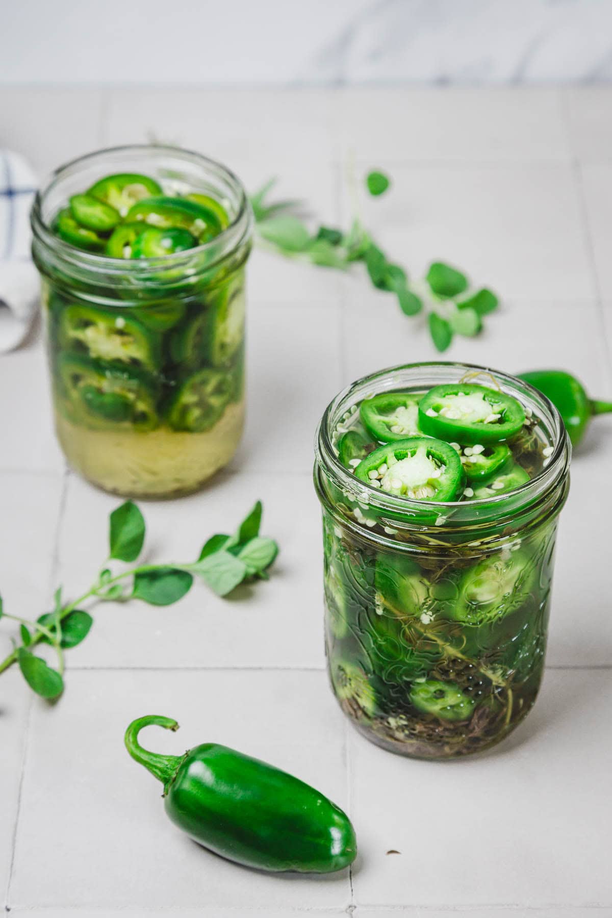 Two glass jars filled with sliced jalape&ntilde;os in brine, with fresh herbs and a whole jalape&ntilde;o nearby.