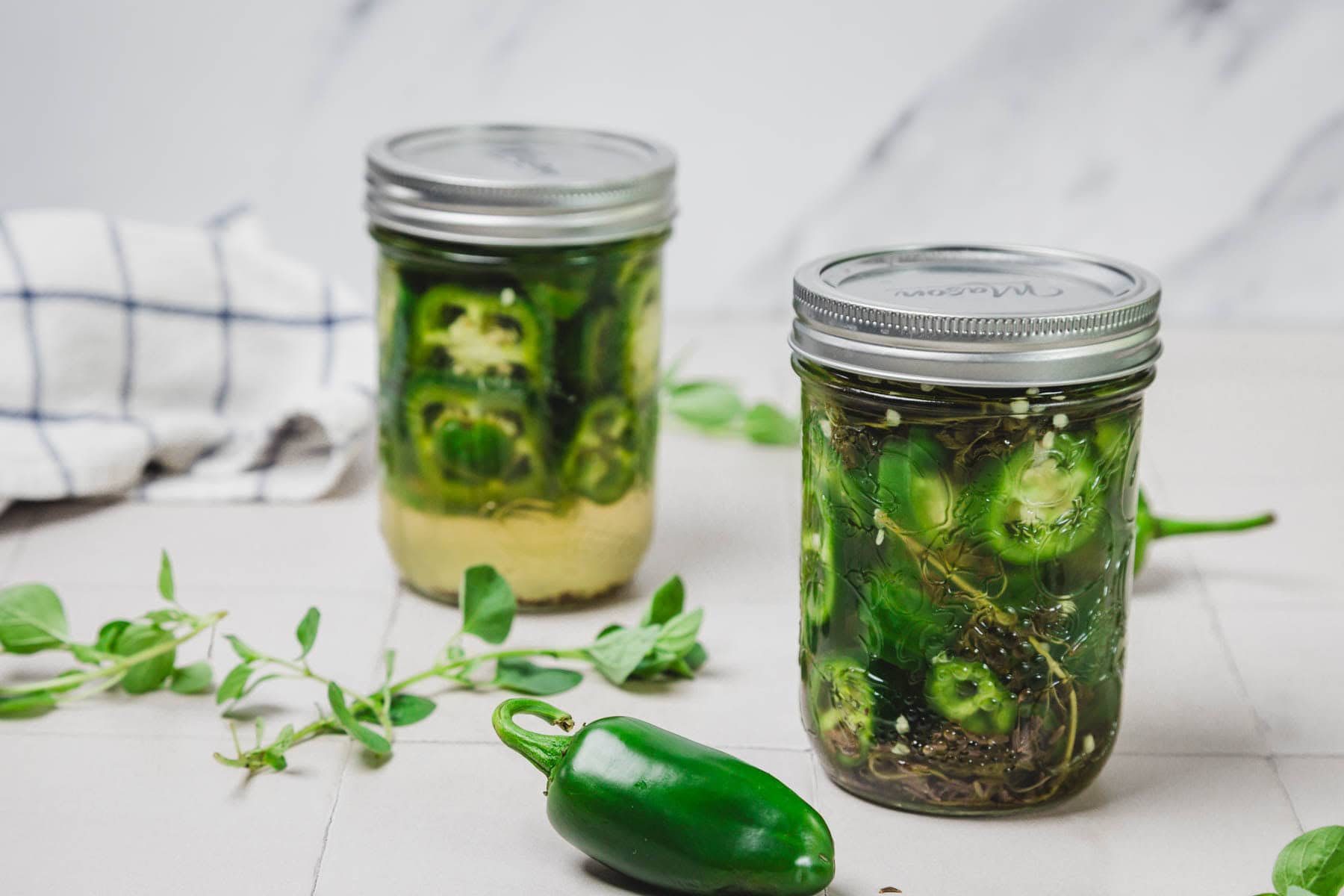 Two jars of pickled jalape&ntilde;os with herbs, a fresh jalape&ntilde;o, and green sprigs on a light surface.