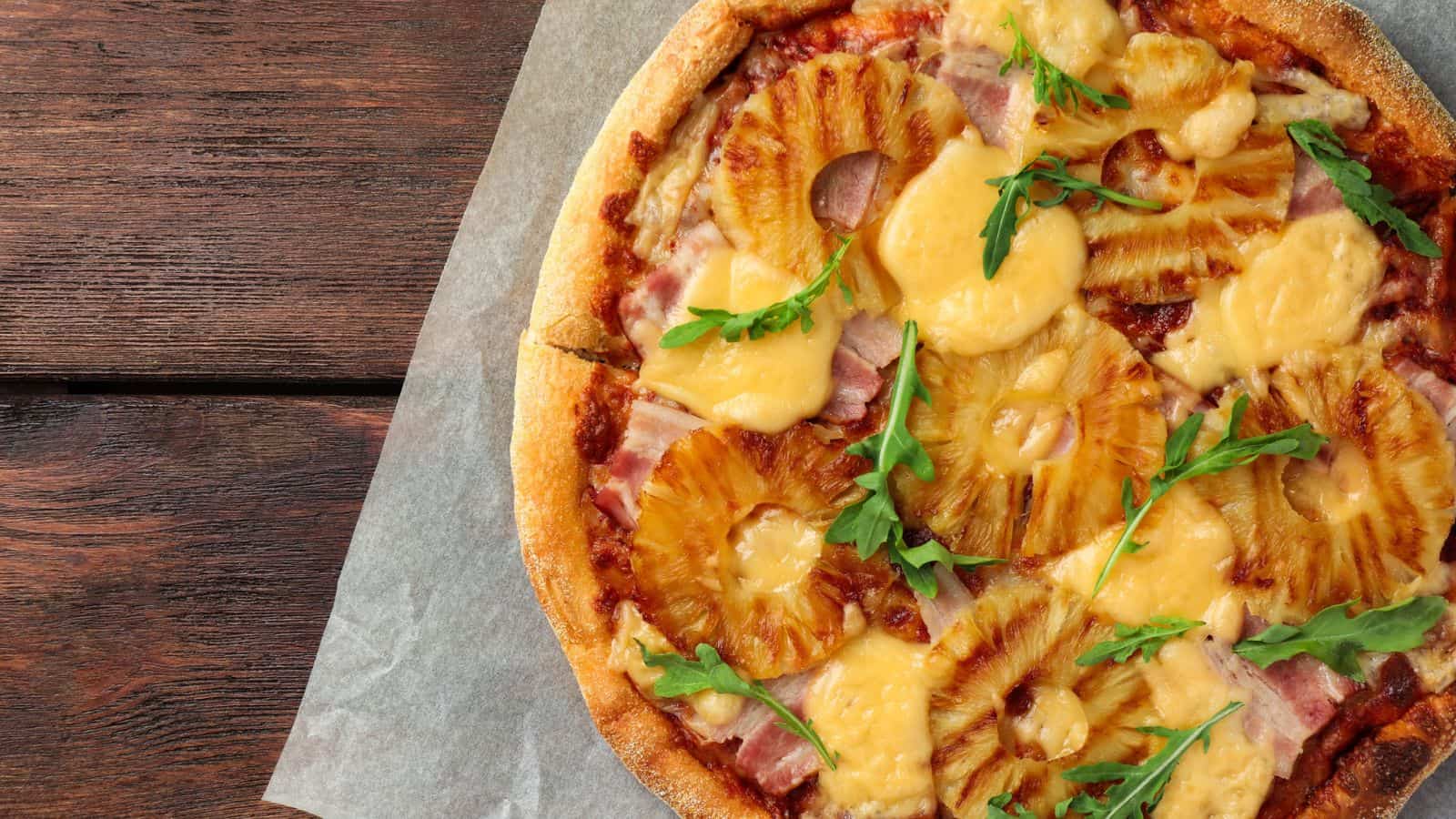 A pizza topped with pineapple rings, melted cheese, ham pieces, and arugula leaves sits on parchment paper next to a wooden surface.