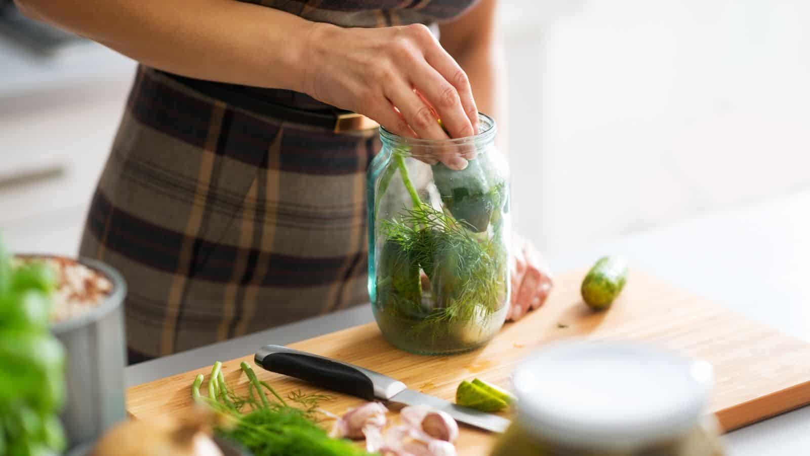 A person placing cucumbers and herbs into a jar for pickling on a kitchen counter, carefully mindful of canning dangers.