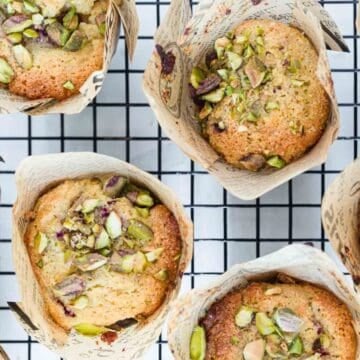 Muffins topped with chopped pistachios in paper wrappers on a cooling rack.