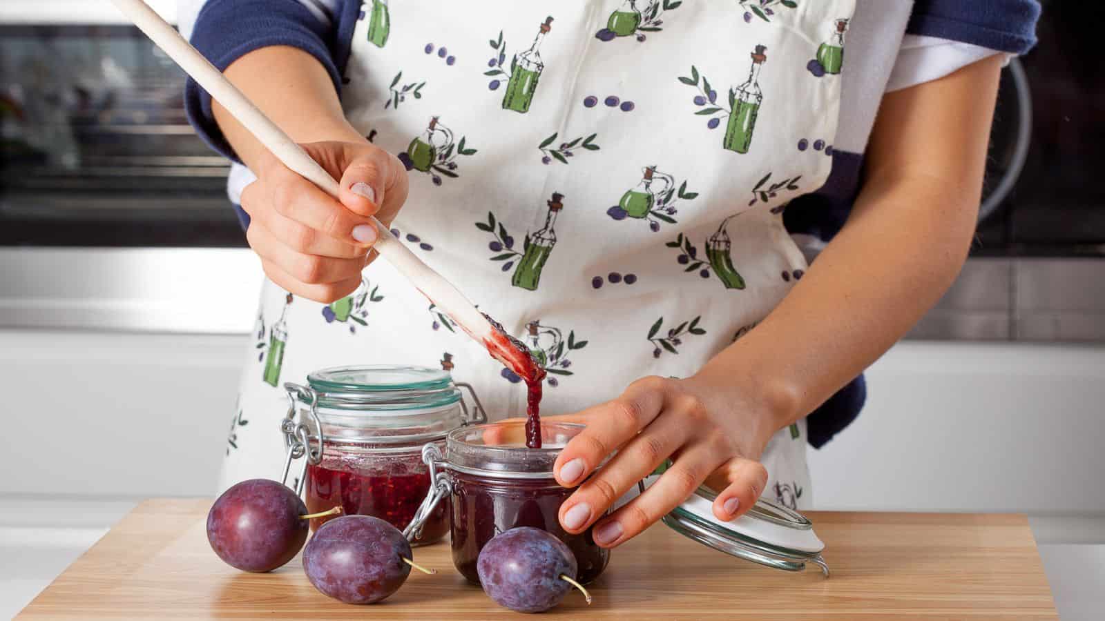 Person filling a glass jar with plum jam using a wooden spoon, with fresh plums on a wooden surface&mdash;always use safe methods to avoid canning dangers.