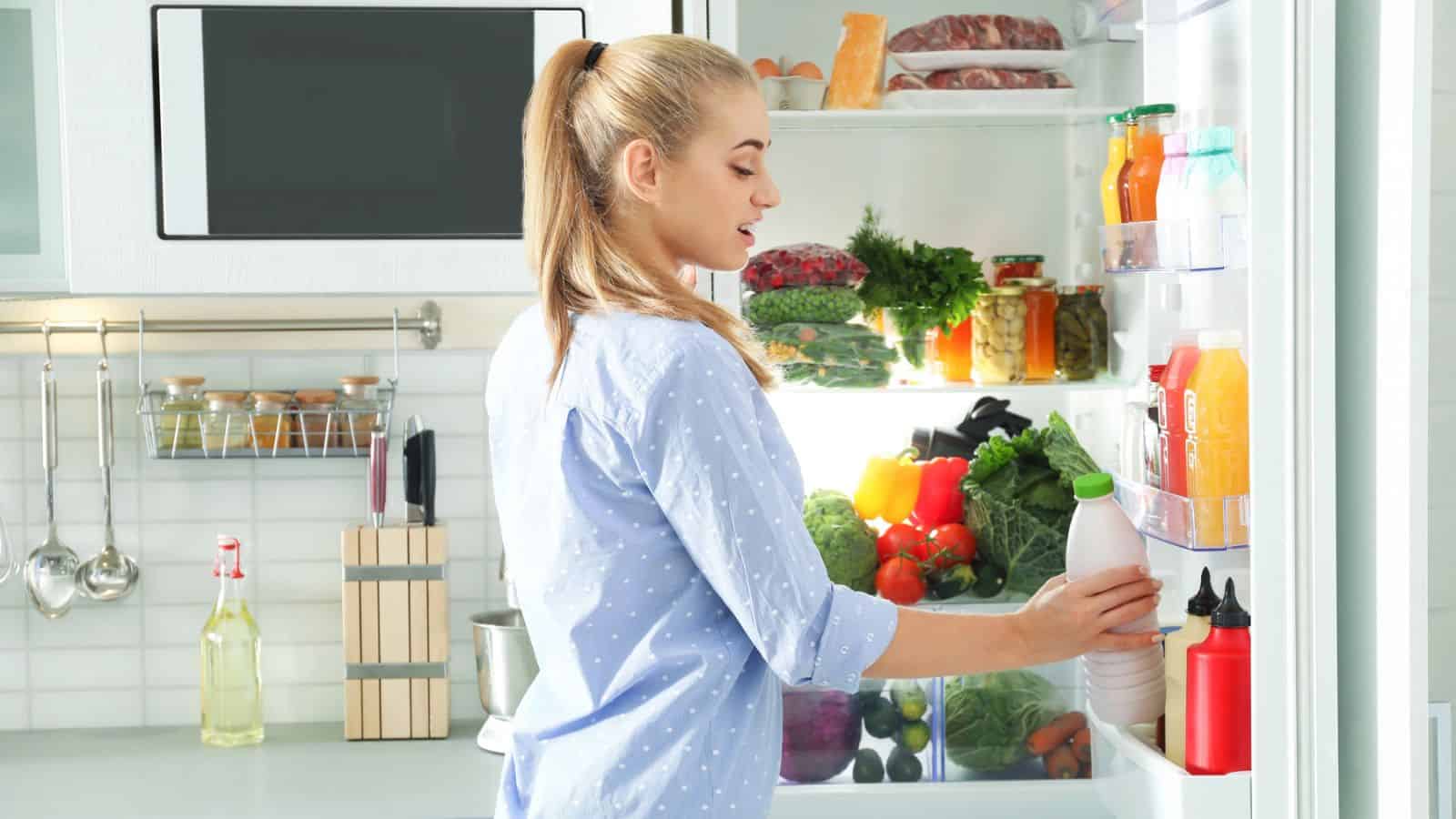 A woman with blonde hair in a ponytail stands in front of an open refrigerator in a modern kitchen, holding a bottle. The fridge is stocked with fresh vegetables, beverages, and other food items.