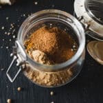 Open glass jar filled with fragrant Shawarma Spice, sitting on a dark surface with scattered spice seeds nearby.